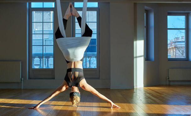 Güzel kız yoga eğitmeni yoga odasında baş aşağı asılı halatlar üzerinde medutiruet gösteriyor. Konsept yoga, esnek vücut, sağlıklı yaşam tarzı, fitness. - Fotoğraf, Görsel