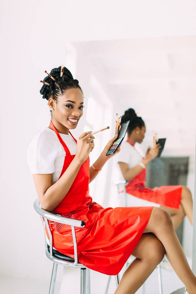 Beautiful young African American beautician woman holding set of make up brushes and watching in the mirror - Foto, afbeelding
