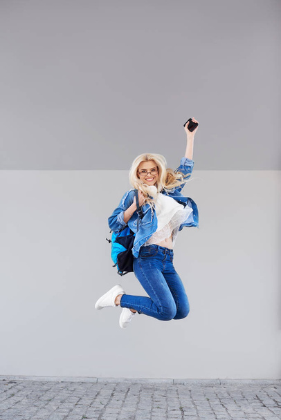 Full body Portrait of happy young woman walking with bag and cellphone. Cheerful student jumping in air over gray background - Foto, afbeelding