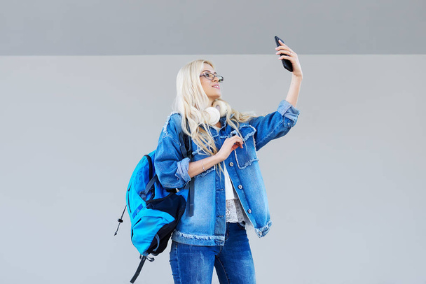 Portrait of a serious and surprised casual girl student with backpack makes a selfie on mobile phone over grey background - Photo, image