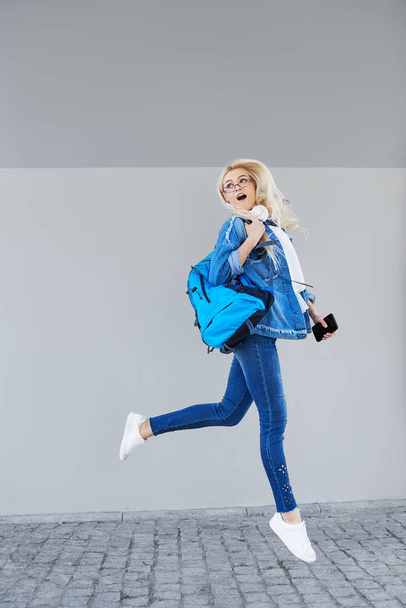 Cuerpo completo Retrato de mujer joven y feliz caminando con bolso y teléfono celular. Estudiante alegre saltando en el aire sobre fondo gris
 - Foto, imagen