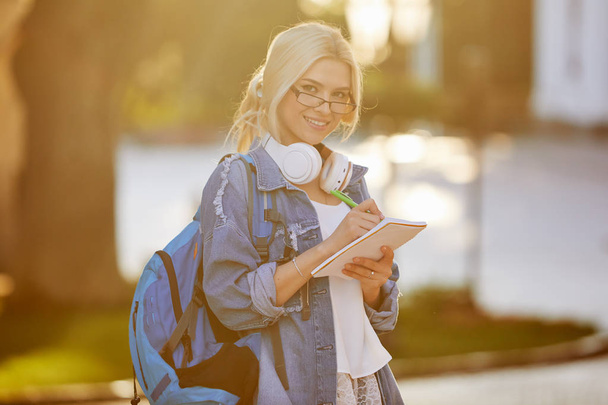 Portret uśmiechniętej młodej kobiety uczeń w okularach z plecaka słuchania muzyki ze słuchawkami i pisania notatek. Koncepcja edukacji online i zdalnej - Zdjęcie, obraz