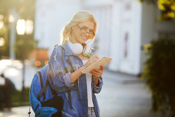 Portrait of a smiling young woman student in eyeglasses with backpack listening music with headphones and writing notes. Online and remote education concept - Фото, изображение