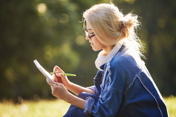 Doğa parkında oturan gözlüklü, gülümseyen bir genç kadın öğrencinin portresi. Kulaklıklarla müzik dinleme ve notlar yazma. Çevrimiçi ve uzaktan eğitim konsepti - Fotoğraf, Görsel