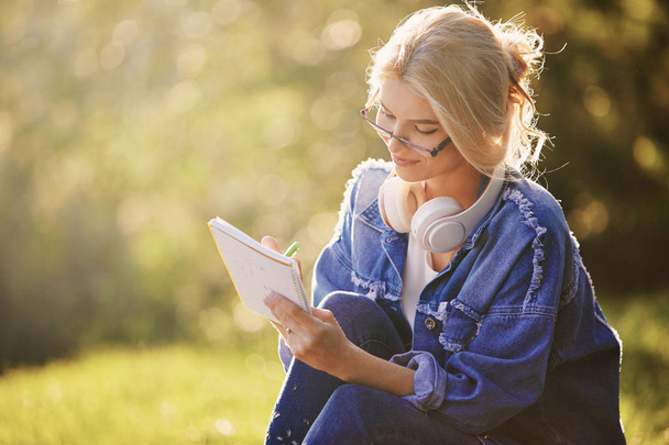 Doğa parkında oturan gözlüklü, gülümseyen bir genç kadın öğrencinin portresi. Kulaklıklarla müzik dinleme ve notlar yazma. Çevrimiçi ve uzaktan eğitim konsepti - Fotoğraf, Görsel