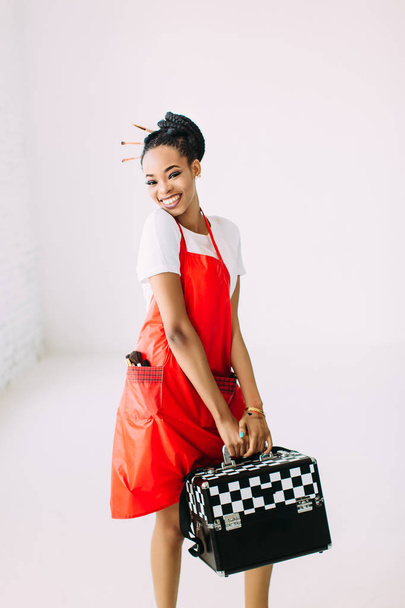 Beautiful young African American beautician woman holding set of makeup equipment in special case, isolated on white background - Фото, зображення
