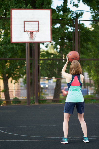 Рыжая тощая девушка бросает мяч в баскетбольное кольцо на открытом воздухе
 - Фото, изображение