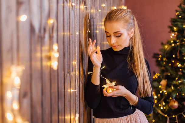 Beautiful young woman dressed in a sweater and skirt standing next to the wooden wall with star and moon lights - Zdjęcie, obraz