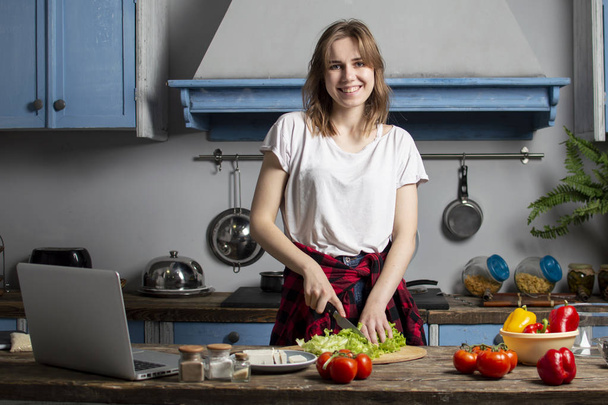 young girl prepares a vegetarian salad in the kitchen and looks into a laptop, she learns to cook by a video tutorial, preparing healthy food - Photo, image