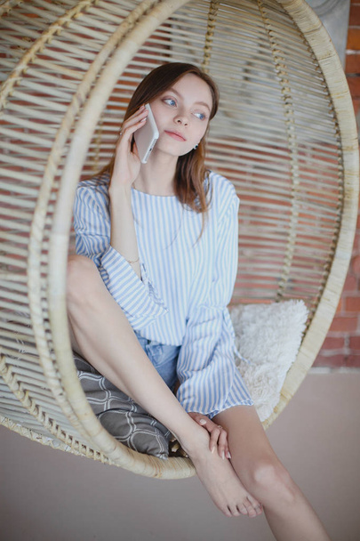 Young pretty girl is sitting in chair and talking on phone - Фото, изображение