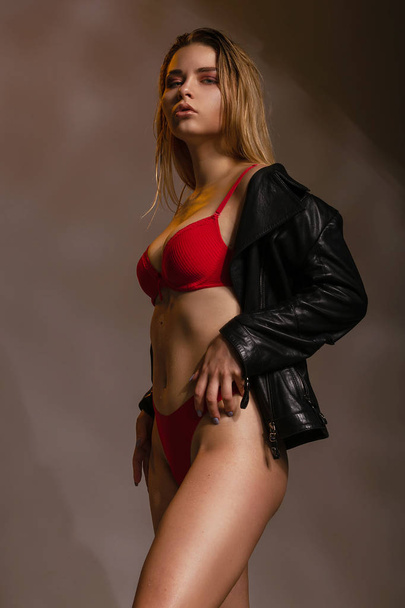 Uwodzicielska Kobieta w skórzanej kurtce i czerwonej bieliźnie erotycznej. Szczupła piękna dziewczyna w seksownej luksusowej bieliźnie z sportowym rysunkiem pozuje na ciemnym szarym tle. - Zdjęcie, obraz