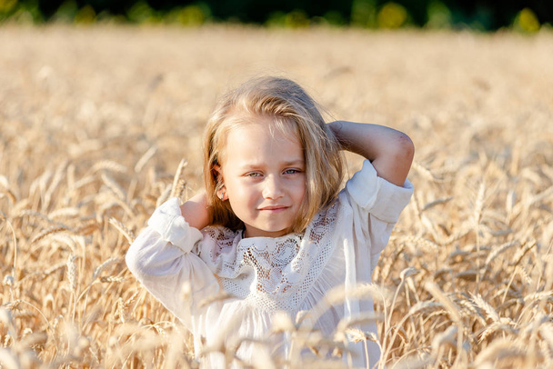 kis szőke lány áll a búzamezőn nyáron és mosolyog - Fotó, kép