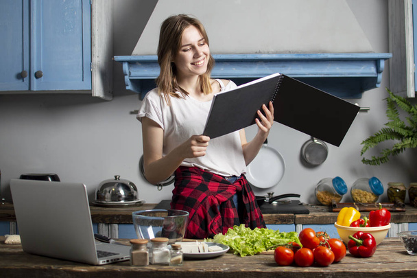 junges Mädchen bereitet in der Küche einen vegetarischen Salat zu und schaut in ein Rezeptbuch, lernt kochen und liest - Foto, Bild