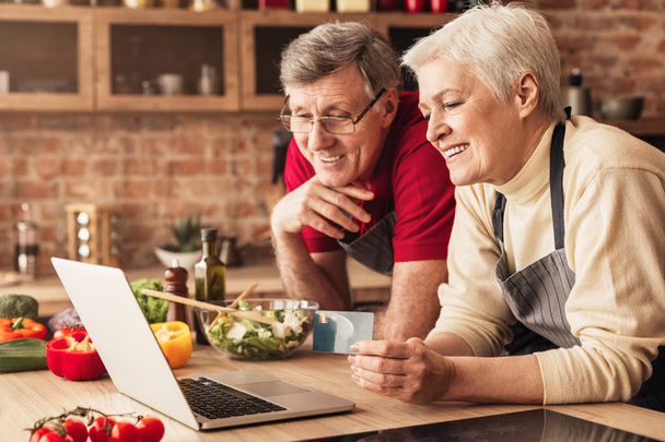 Happy seniorit tilaamalla illallisen verkossa kannettavan tietokoneen ja luottokortin keittiössä
 - Valokuva, kuva