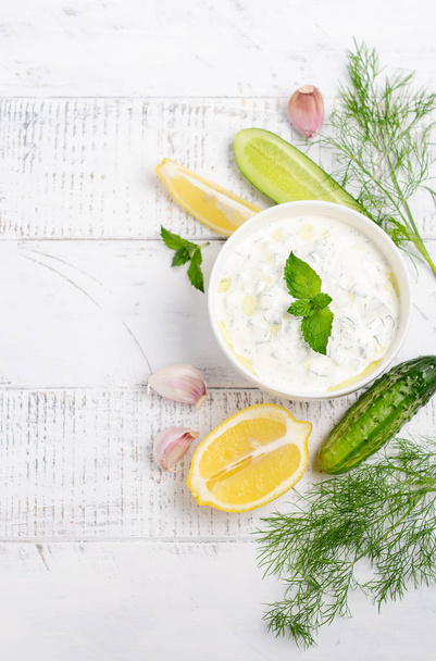 Greek dip sauce or dressing tzatziki and ingredients decorated with olive oil and mint on white wooden table  - Valokuva, kuva