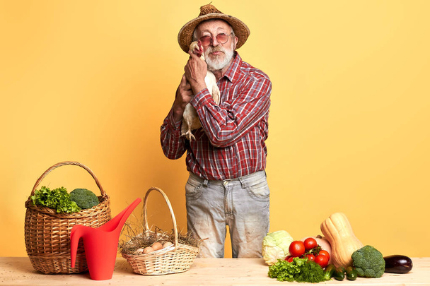Senior gardener, posing with hen in hands, standing behind counter with vegetables - Fotó, kép