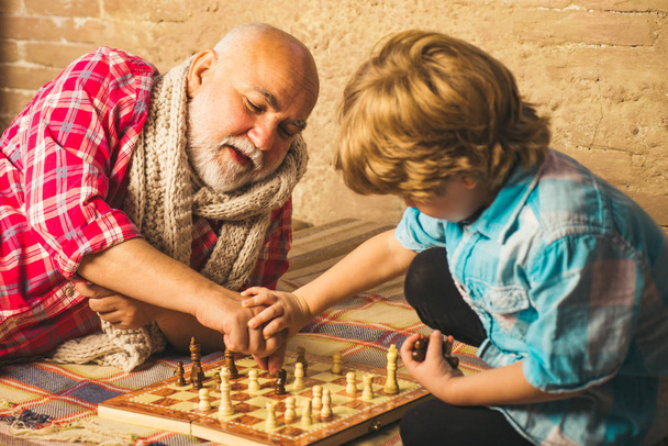 Handsome grandpa and grandson are playing chess while spending time together at home. Chess hobbies - granddad with grandson on a playing chess. - Fotó, kép