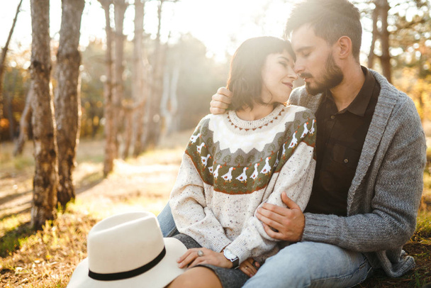 Lovely hipster couple looking at each other. Couple wearing beautiful hats and sweaters. Lifestyle, happy couple of two play on a sunny day in the park. The concept of youth, love and lifestyle. Sunset in autumn  - 写真・画像