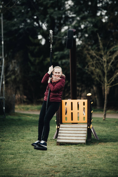 Young woman sitting on a swing in park. Pretty girl swinging on a swing against the urban landscape. Female feeling happy and healthy having fun in a sunny beautiful day - Photo, Image