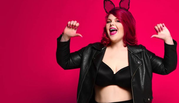 Cute sexy plus size brunette with black bunny ears in leather jacket and underwear posing on red background - Фото, изображение