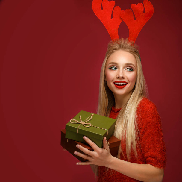 Hermosa chica rubia en una imagen de Año Nuevo con cajas de regalos en las manos y cuernos de ciervo en la cabeza. Cara de belleza con maquillaje festivo
. - Foto, Imagen