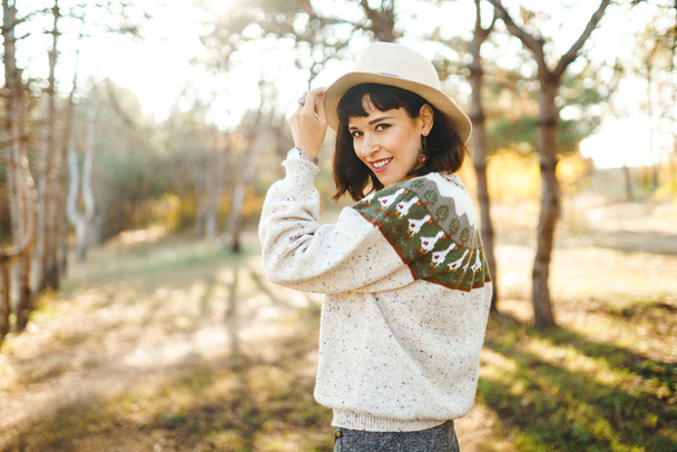 Lovely girl with a beautiful smile at sunset in the forest. The girl is dressed with a hat, a light sweater and a skirt. Knee-high boots. Hipster girl concept, lifestyle style, autumn style. - Zdjęcie, obraz
