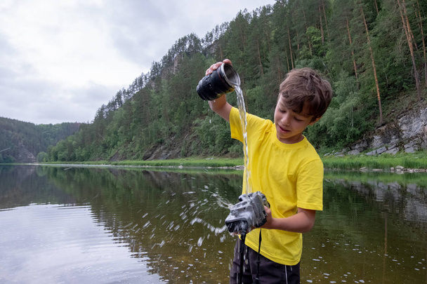 Doğada bir dağ nehrinde çılgın temizlik fotoğraf ekipmanları. Sarı tişörtlü bir çocuk elinde lens olmadan bir kamera tutar ve içine su döker. - Fotoğraf, Görsel