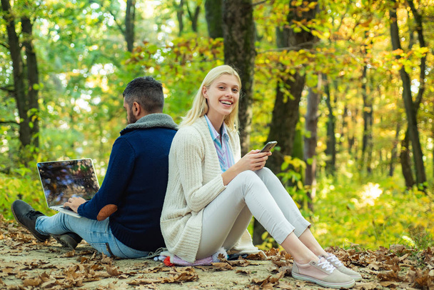Happy couple on autumn walk. Business couple - man browsing Internet on a notebook, woman using mobile phone outdoors. Business outdoor in autumn park. - 写真・画像