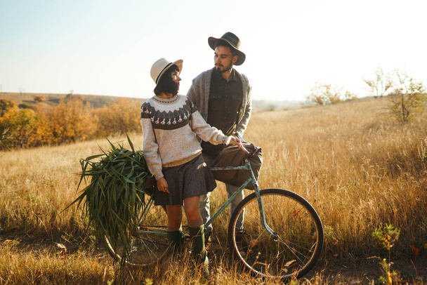 Lovely hipster couple with bike. Couple wearing beautiful hats and sweaters. Lifestyle, happy couple of two play on a sunny day in the park. The concept of youth, love and lifestyle. Sunset in autumn - Photo, image
