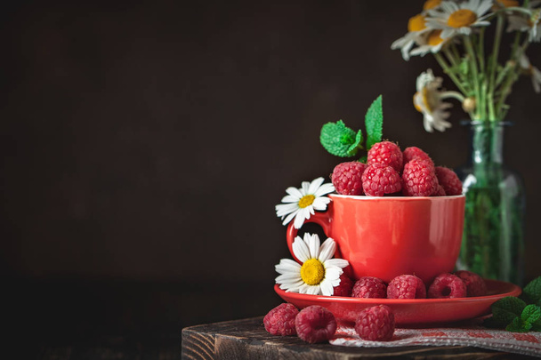 Raspberry in a red cup with chamomile and leaves on a dark background. Summer and healthy food concept. Background with copy space. Selective focus. - Foto, immagini