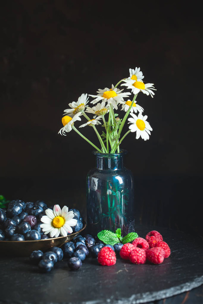 Raspberries and blueberries with chamomile and leaves on a dark background. Summer and healthy food concept. Background with copy space. Vertical. - Photo, image
