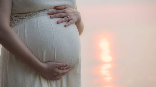 Μια έγκυος γυναίκα στέκεται κοντά στη θάλασσα στο πίσω μέρος του κρεβατιού της, σε κοντινό πλάνο, χαϊδεύοντας την κοιλιά της - Φωτογραφία, εικόνα