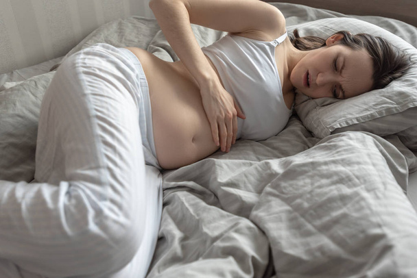 Nuori raskaana oleva nainen makaa sängyssä ja pitää vatsa
 - Valokuva, kuva