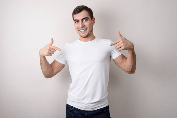 portrait of smiling handsome young man in a white blank t-shirt shows her with his fingers, place for design - Photo, Image