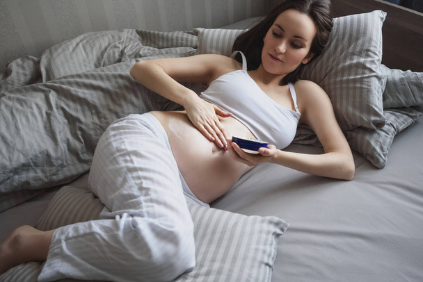 Jonge Europese zwangere vrouw liggend in bed en masserende buik crème, huidverzorging - Foto, afbeelding