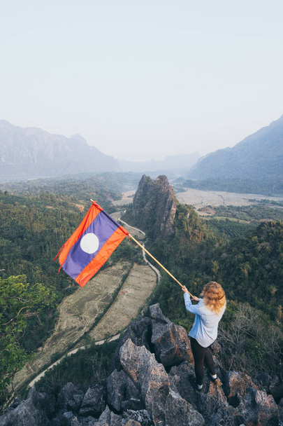 Donna bionda in piedi sulla vetta della montagna con la bandiera laotiana in mano che domina la valle di Vang Vieng, Laos
 - Foto, immagini