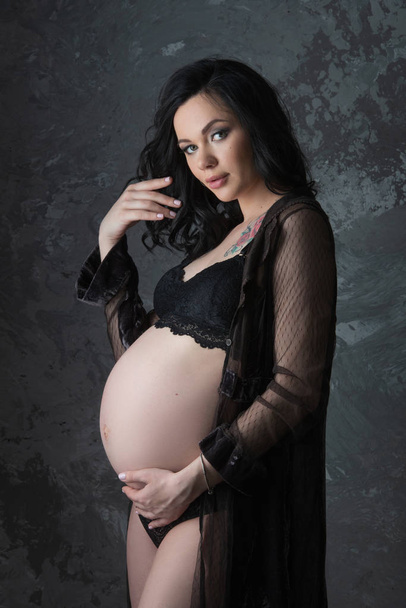 The concept of pregnancy. Beautiful pregnant woman in black underwear hugging her tummy against a dark background - Fotó, kép