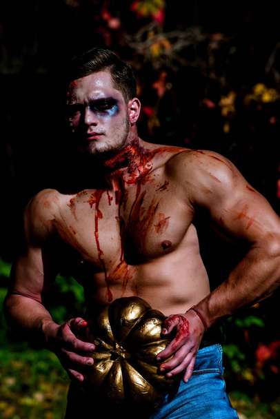 Halloween face art. Young handsome man with muscular body posing in street holding halloween pumpkin. Big zombie man halloween vampire or bloody war soldier with wounds and red blood with pumpkin. - Photo, Image