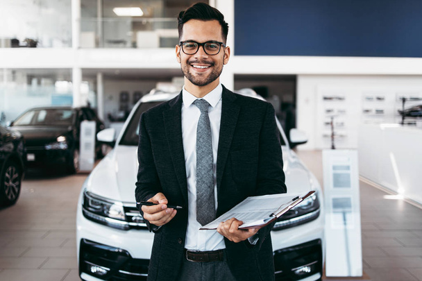 Good looking, cheerful and friendly salesman poses in a car salon or showroom. - Fotó, kép