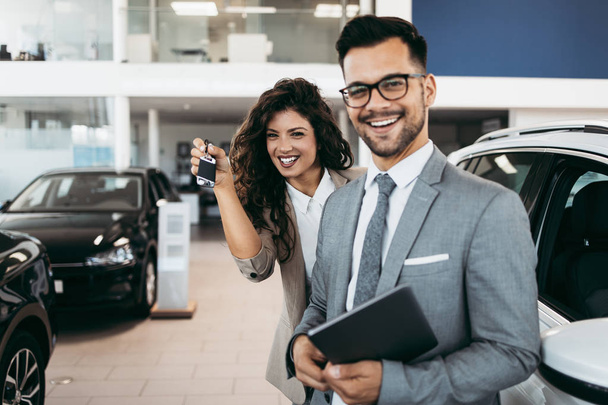 Happy and beautiful middle age business woman buying new car at showroom. She standing behind nice and positive seller and showing her new car keys. - Φωτογραφία, εικόνα