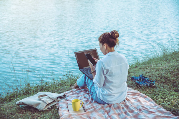 Mulheres sentadas no lado relaxante do rio no parque. segurando o telefone celular e está trabalhando com um notebook e beber café. No parque no feriado
. - Foto, Imagem