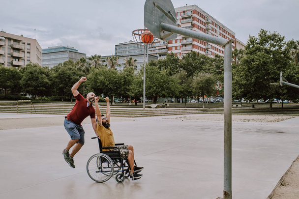 toimintarajoitteinen mies pyörätuolissa pelaa koria ystävän kanssa
 - Valokuva, kuva