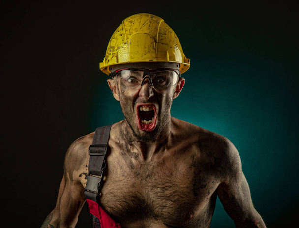 Een man mijnwerker in een werknemer s kleren en een helm, vies, in roet, met een naakte lichaam, grimaces op de camera - Foto, afbeelding