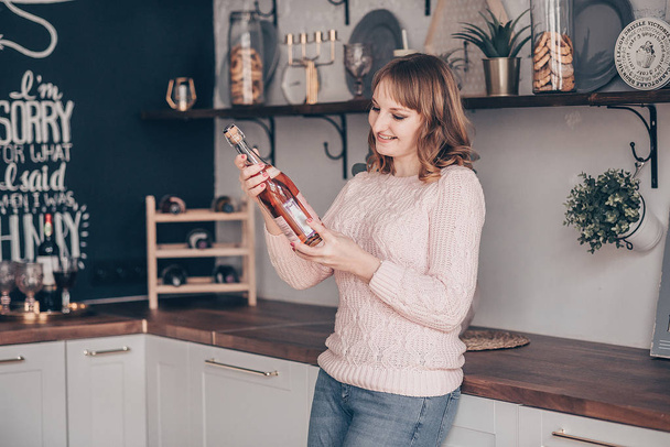 Attractive young female with curly hair. Holds bottle of alcoholic drink, reads information about wine, looks happy. Beautiful girl finds champagne. Woman chooses wine in the kitchen. - Fotoğraf, Görsel