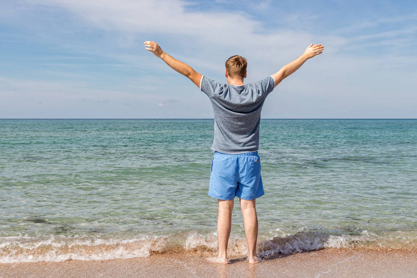 Young man in a t-shirt on the seashore and looks at the sea, back view - Foto, imagen