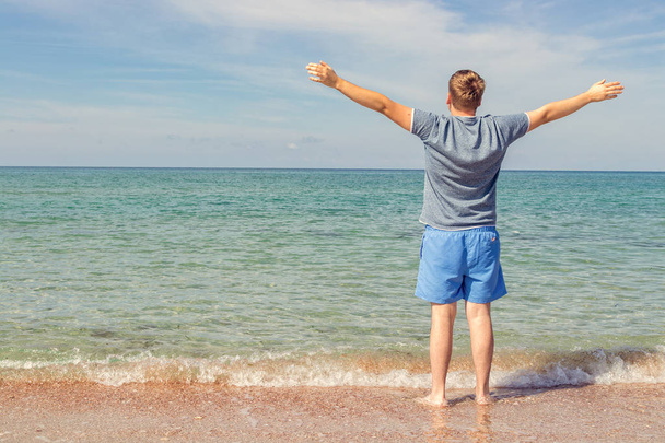 Młody człowiek w t-shirt na brzegu morza i patrzy na morze, widok z tyłu - Zdjęcie, obraz