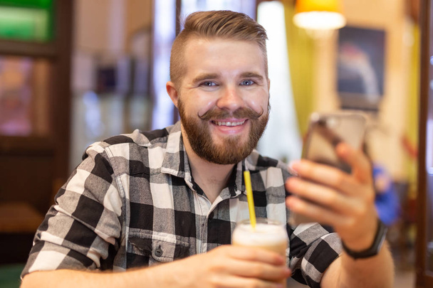 Χαριτωμένο θετικό νεαρό χίπστερ με μουστάκι και γενειάδα να χαμογελάει ποζάρισμα σε μια καφετέρια. Η έννοια των αστείοι χαρούμενοι άνδρες. - Φωτογραφία, εικόνα