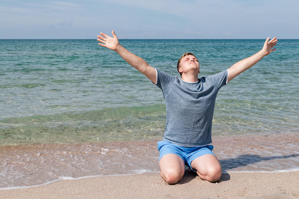 Счастливый молодой человек в футболке сидит на берегу моря и смотрит на море, вид сзади
 - Фото, изображение