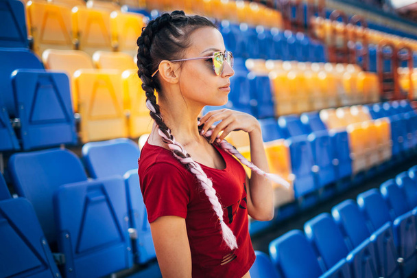 Young beautiful woman teen girl listen music outdoor earphones headphone on smartphone lying on the grass sitting on sport stadium attractive braided pink hair braids bubble gum summer hot pink glasses eating ice cream - Фото, зображення
