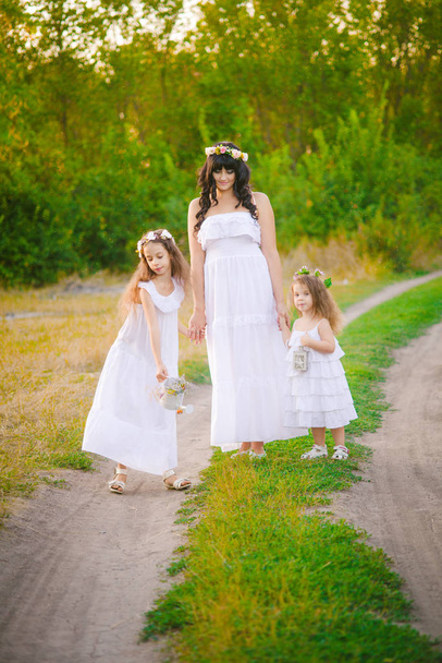 Young mother with her daughters in white dresses having fun in a summer field at sunset by the river - Photo, image